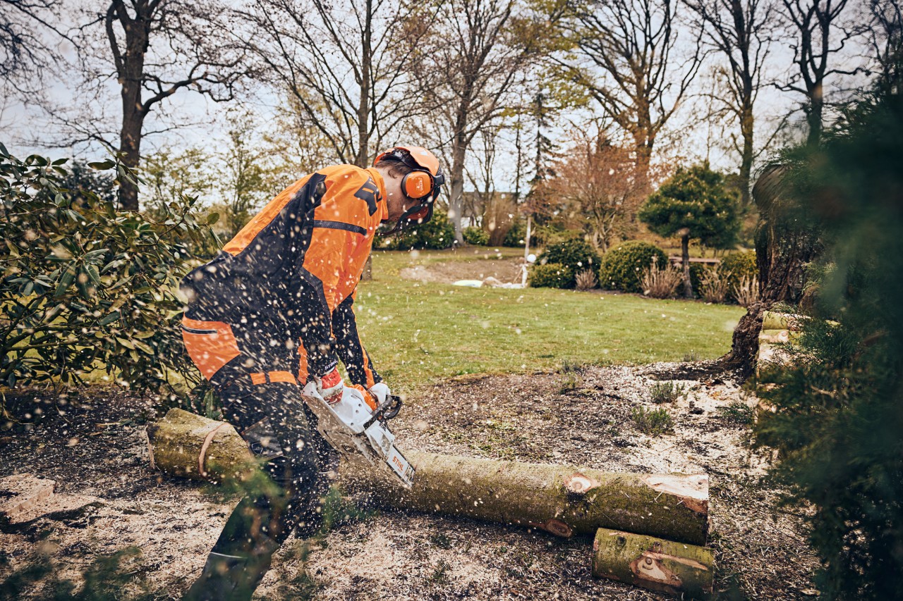 Mężczyzna w wyposażeniu ochronnym tnie pień drzewa na terenie zielonym za pomocą akumulatorowej pilarki łańcuchowej STIHL