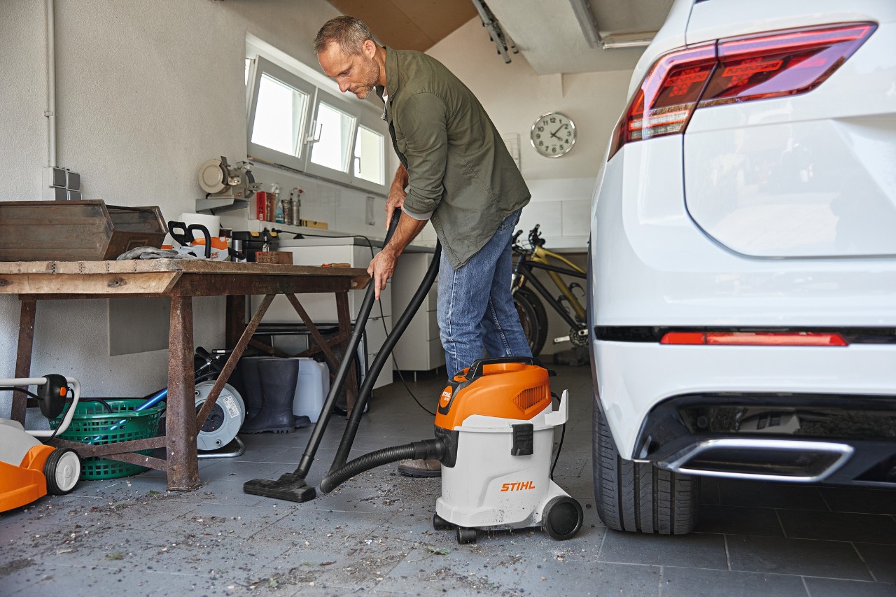 Mężczyzna odkurzający podłogę w garażu za pomocą odkurzacza na mokro i sucho STIHL, obok niego ławka i biały samochód